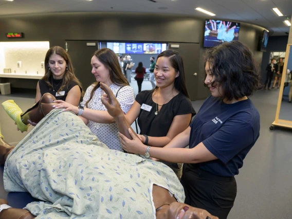 Several students stand alongside a simulation manikin dressed in a hospital gown. 
