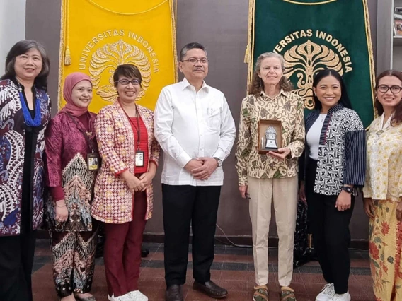 Photo of a row of seven people in front of two banners reading “Universitas Indonesia.” Priscilla Magrath, third from the right, holds a plaque in one hand. 