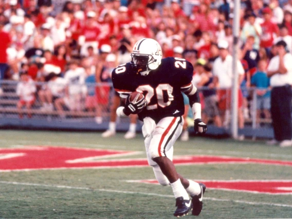 An action photo of a football player wearing a white helmet, blue jersey and white pants trimmed in red and blue while running a football.