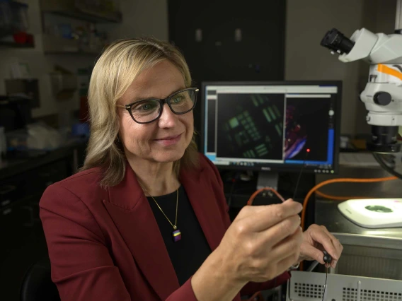 Jennifer Barton, PhD, wearing a red blazer and glasses is working in a laboratory, holding a small object. A computer screen displaying a microscopic image is visible in the background, along with a microscope, other lab equipment and shelves.