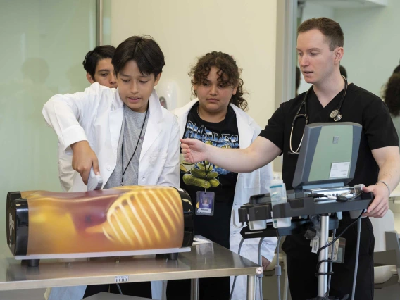 A 13-year-old boy holds a medical tool against a model of a torso while a medical student in scrubs and two other middle schoolers watch.