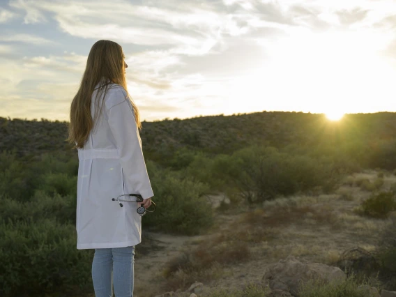 A woman wearing a white medical coat holds a stethoscope while looking towards a sunset over a rural desert landscape.