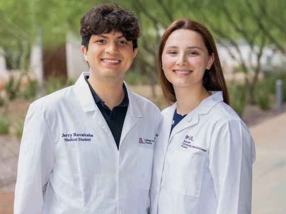 Medical students Jerry Ruvalcaba and Katie Simic stand side by side outdoors, smiling in their white lab coats embroidered with titles from the University of Arizona College of Medicine - Phoenix.