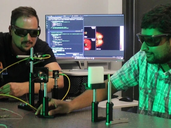 Two male researchers wearing dark safety glasses test a prototype of an imagine device in a lab. Behind them is a computer screen that shows an image of the sample being studied.
