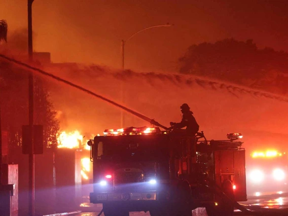 A nighttime photo shows a firefighter standing on a firetruck and shooting water toward burning buildings during the January 2025 Palisades Fire in California.