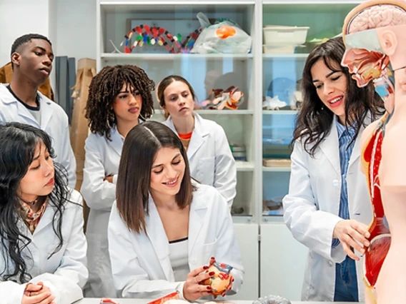 A nursing instructor stands next to a manikin and points out parts of its body as she instructs five students. All the students and the instructor are wearing white lab coats.