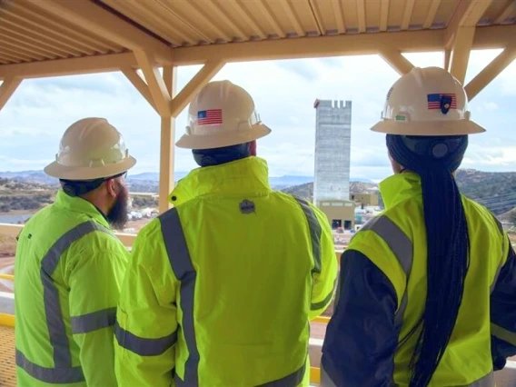Two men and a woman, all dressed in hard hats and reflective mining jackets, looks at a mining facility in the distance. Their backs are to the camera.
