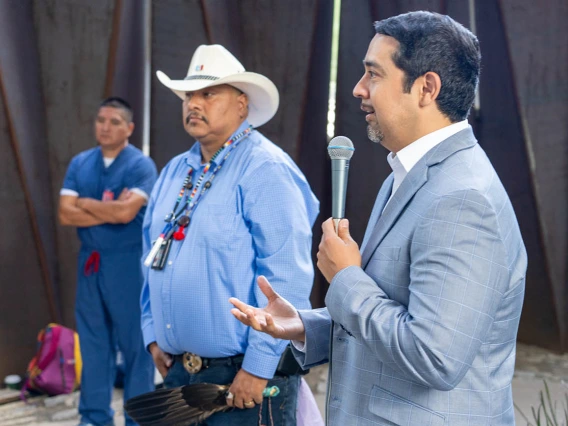 A man in a gray suite holding a microphone talks in an outdoor setting with a man in a straw cowboy had and beeded necklace stands in the background. 