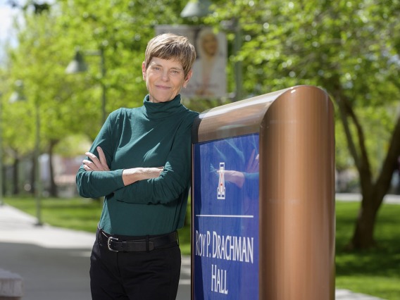 Woman with short brown hair wearing a green turtleneck and black pants leans again the University of Arizona Roy P. Drachman Hall sign