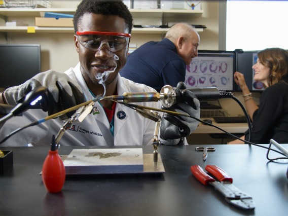 Ike Chinyere solders wiring for a cardiac electrophysiology model to study heart arrhythmias. Steven Goldman, MD, and Elizabeth Juneman, MD, research mentors, in background.