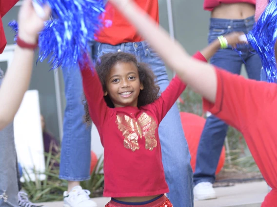 A group of people of mixed ages all wearing red and blue are smiling and dancing waiving pom poms.