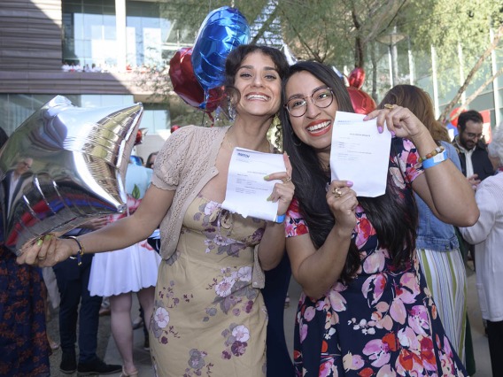 Two women in dresses with long dark hair hold up match day letters and smile big