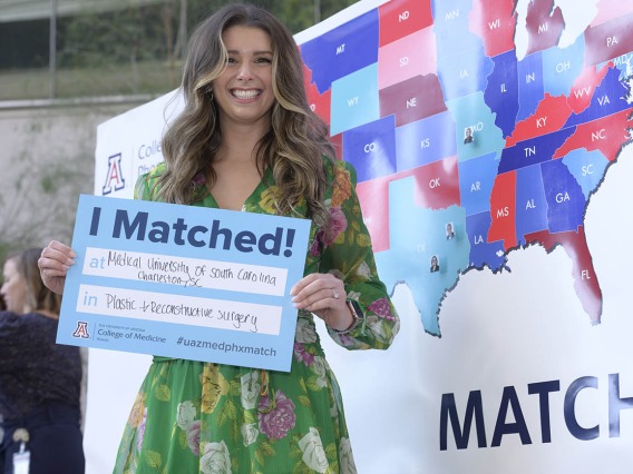 Young woman in dress and long brown hair holds a poster that says "I matched" in front of a map of the U.S.
