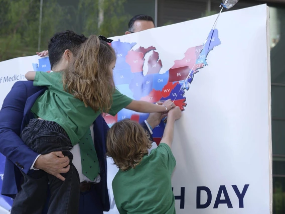 A man in a blue suit holding a girl and next to a boy stick a pin in a map of the U.S.