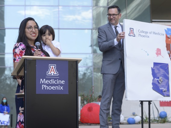 A young adult woman with long dark hair stands at a UArizona podium holding a child. 