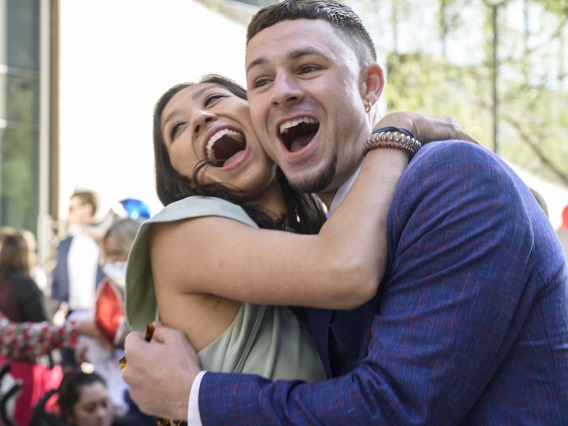 A young woman and man with light skin hug each other with huge open-mouthed smiles.