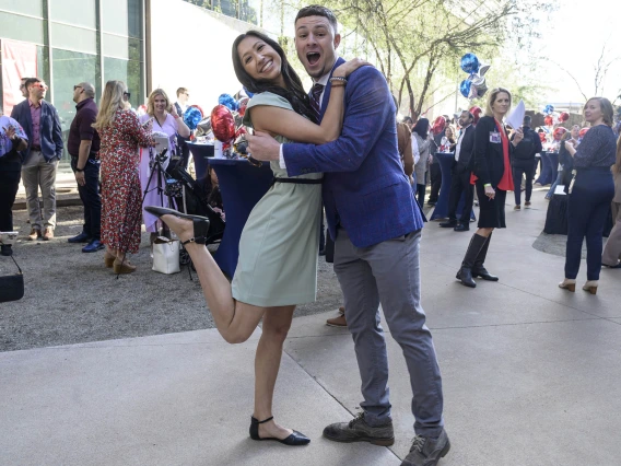 Young adult man and woman embrace, looking at viewer smiling and woman has her leg bent up behind her. 