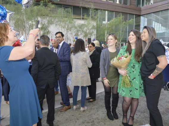 A woman with red hair takes a photo of three woman standing next to each other smiling. The woman in the middle is holder flowers. 