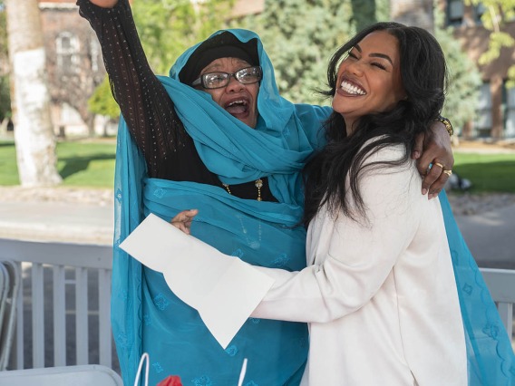 An older dark-skinned woman smiles and holds up one arm in celebration as she hugs a smiling younger dark-skinned woman. 