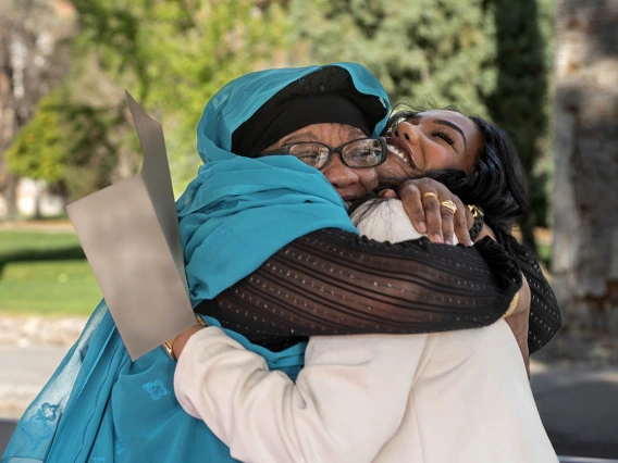 A young woman is hugged by her mother as they both smile. Both have dark skin and the young woman is holding a letter. 