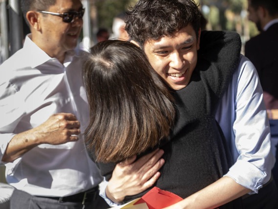 A young man with wiht a big smile and curly dark hair hugs his mom.