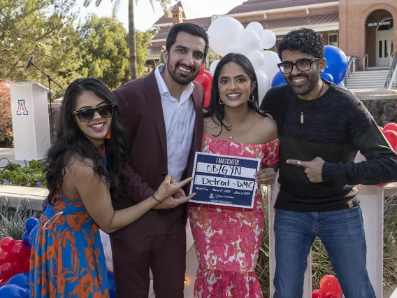 Four young adults, two men and two women, with dark skin and hair smile while the woman in the center holds a sign signifying where she matched.