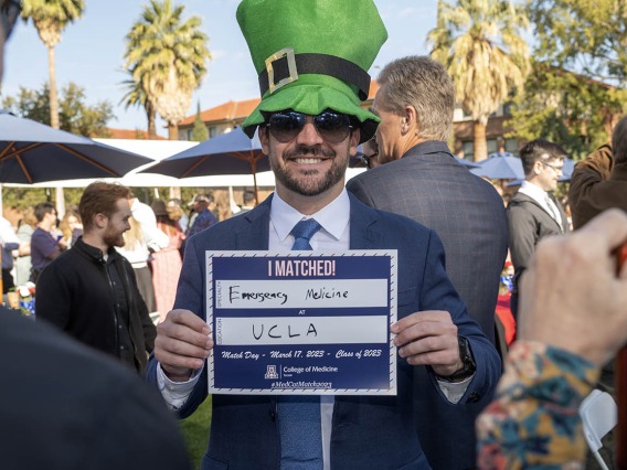 A man with a beard wearing a suite and big green hat hods a sign that says "I matched at UCLA"