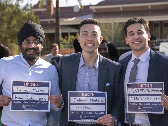 Three young adult men, all smiling, stand in a row holding signs stating where they were matched. 