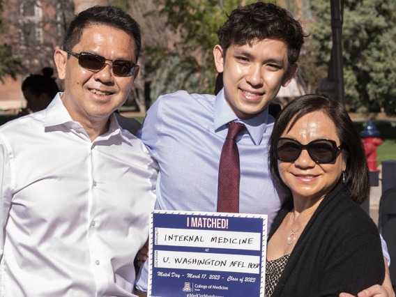 A young Asian man with a dress shirt and tie poses between his mom and dad. They are holding up a sign saying where he matched. 