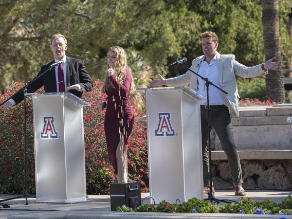 Two men and a woman stand on a stage with two podiums with UArizona logo. Man on right has his hands outstreached and is smiling. 