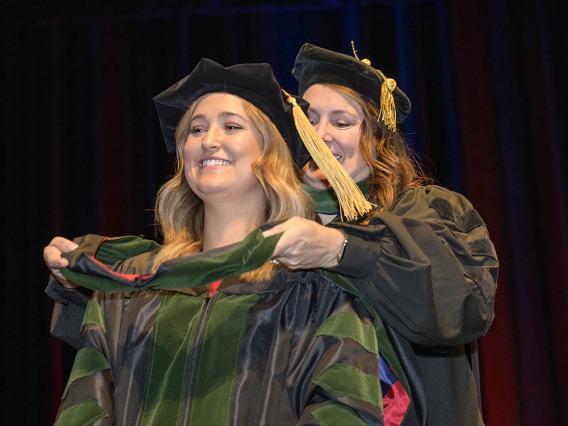 A young blonde woman in graduation regalia smiles as a professor places a sash over her shoulders. 