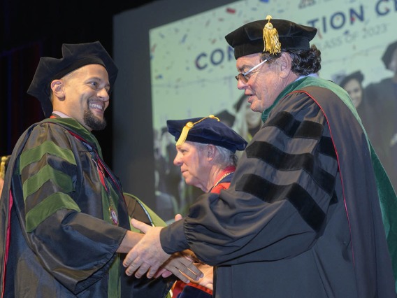 A smiling young man in graduation regalia shakes hands with the dean of the college. 