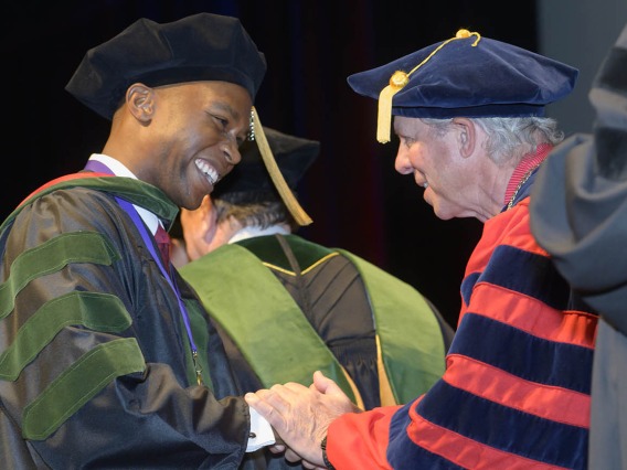 A young man in graduation cap and gown smiles as he shakes hands with the president of the university, also in graduation regalia.