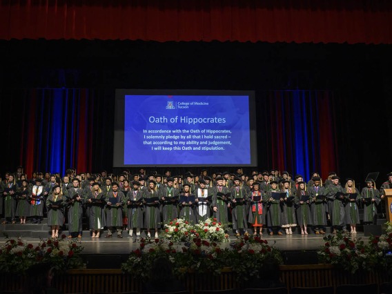 Over 100 medical school graduates in caps and gowns stand on a stage under a large blue display screen with an oath on it. 