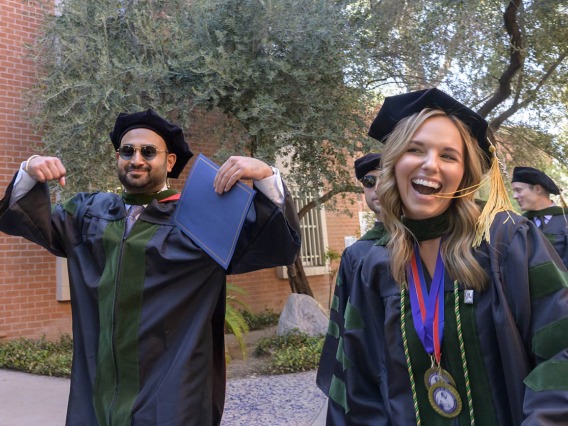 A young man and woman in graduation regalia smile and gesture in celebration as they walk outside. 