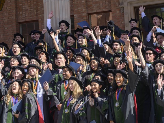 Large group of medical students in graduation robes and hats wave and cheer. 