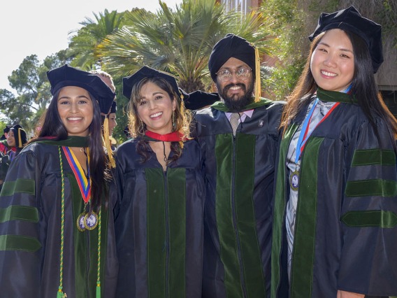 Four medical school graduates in caps and gowns stand together smiling. 