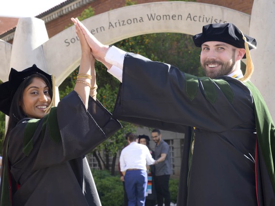 A young woman and man in graduation robes and hats smile as they hold their hands together overhead to make an arch. 
