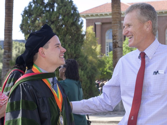 A young male graduate wearing a cap and gown faces an older man who is patting him on the arm in congratulations. 