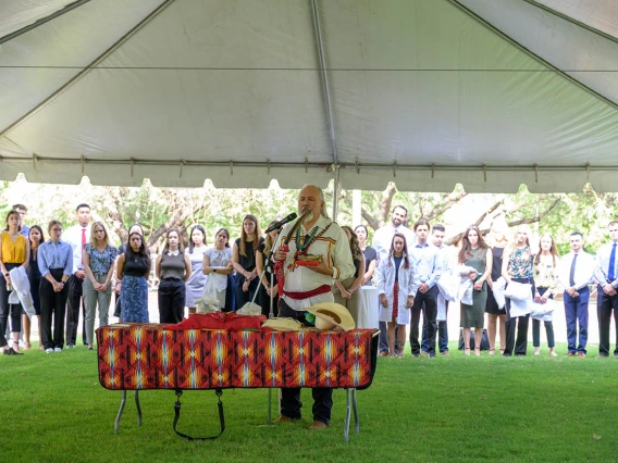 An oudoor photo of a Native American man standing at a table leading a blessing ceremony with a croud of people standing around him. 