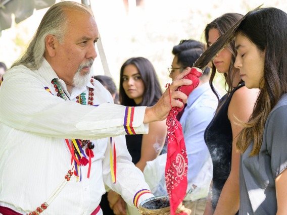 An older man in a traditional Native American designed shirt touches the forehead of a young Native American woman with an eagle feather. 