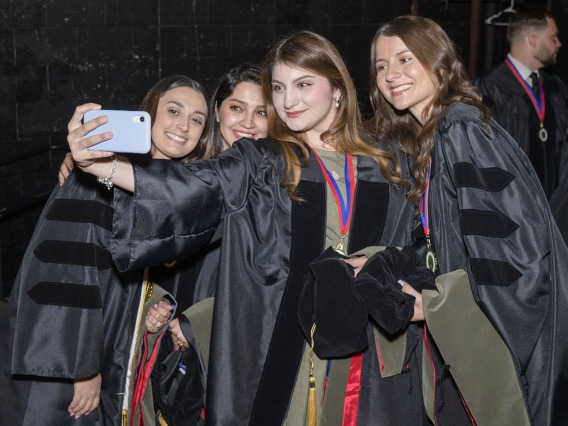 Four young women in black graduation caps and gowns smile as they take a selfie.