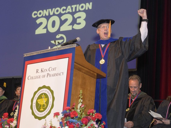 Older white man dressed in graduation cap and gown stands at a podium witih fist in air as if cheering. 