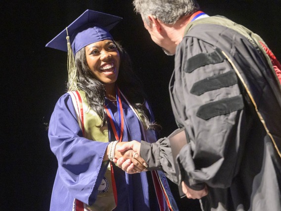 A young Black woman in a blue graduation cap and gown smiles as she shakes hands with a professor. 