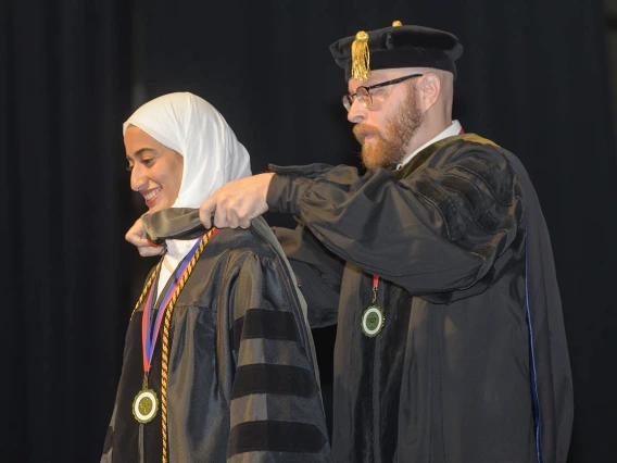 A young woman wearing a white head scarf and a graduation gown has a sash placed over her shoulders by professor.