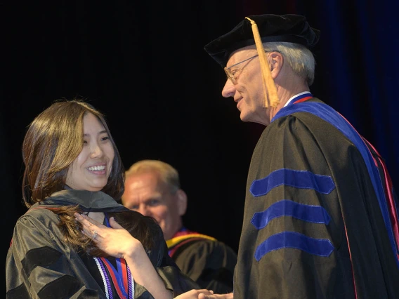 A young Asian woman in a graduation gown shakes hands with an older white man, also in cap and gown. 