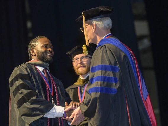 A young Black man in a graduation gown shakes hands with the college dean, an older white man.