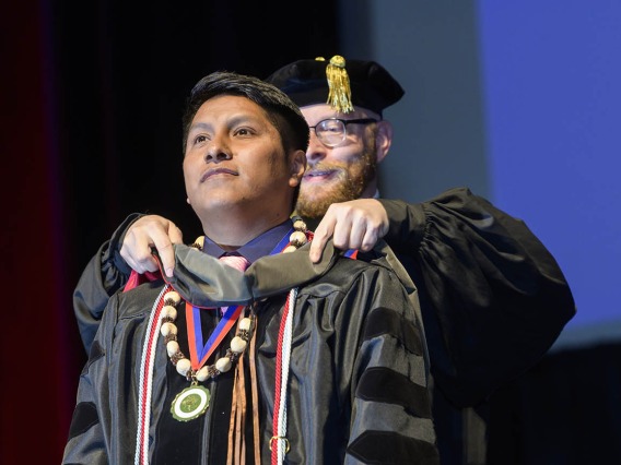 A young Native American man in a graduation gown and beaded necklace has a sash placed over his shoulders by a professor. 