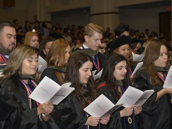 A large group of Pharmacy graduates in graduation gowns read from pamphlets they are all holding. 