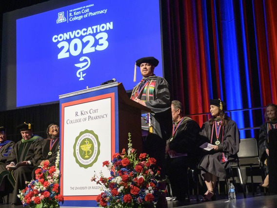 A young man in a black graduation cap and gown stands on a stage at a podium with a screen behind him that says "Convocation 2023"